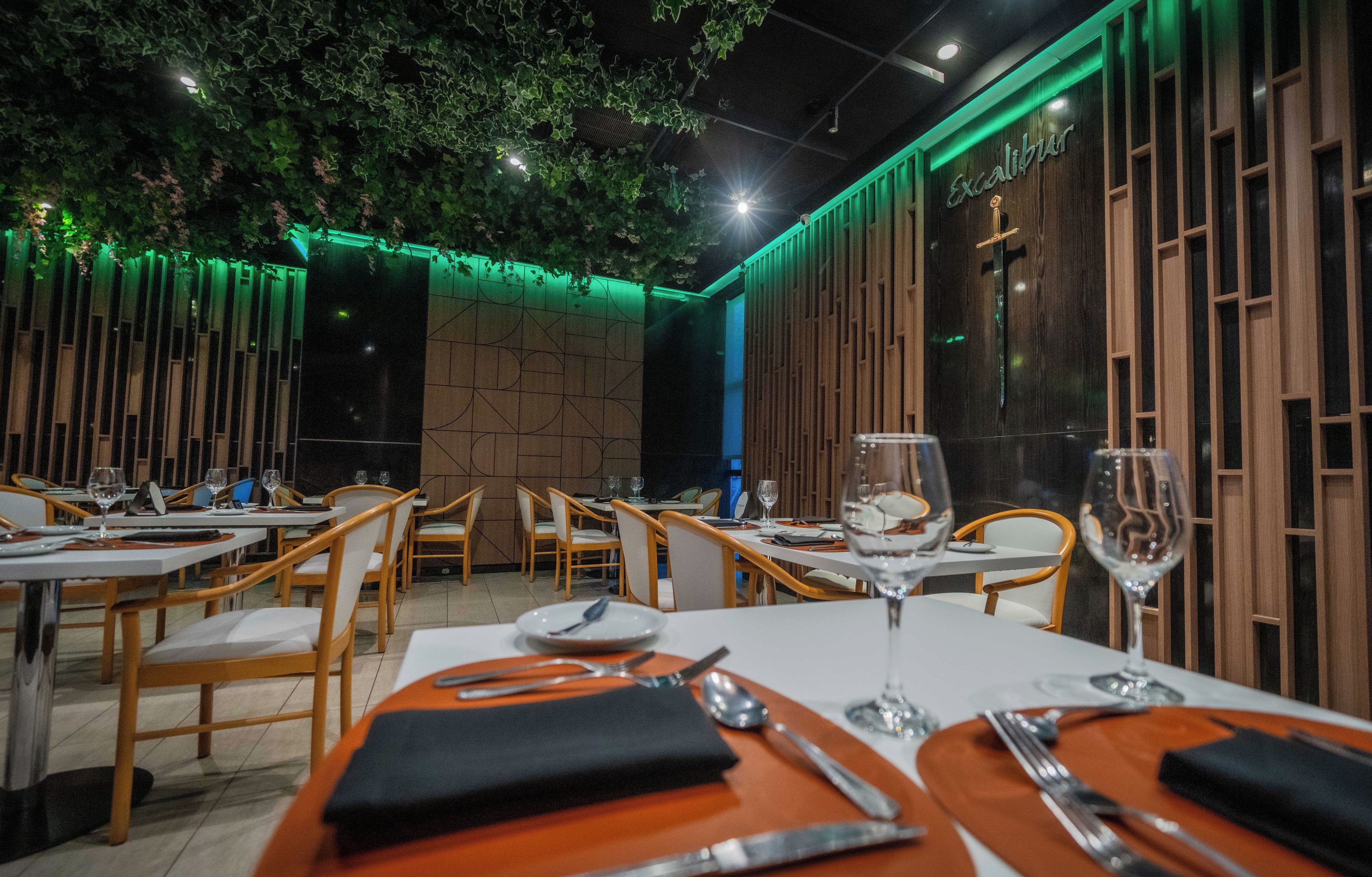 Un restaurante con sillas de madera, mesas dispuestas con cubiertos y una pared iluminada en verde.