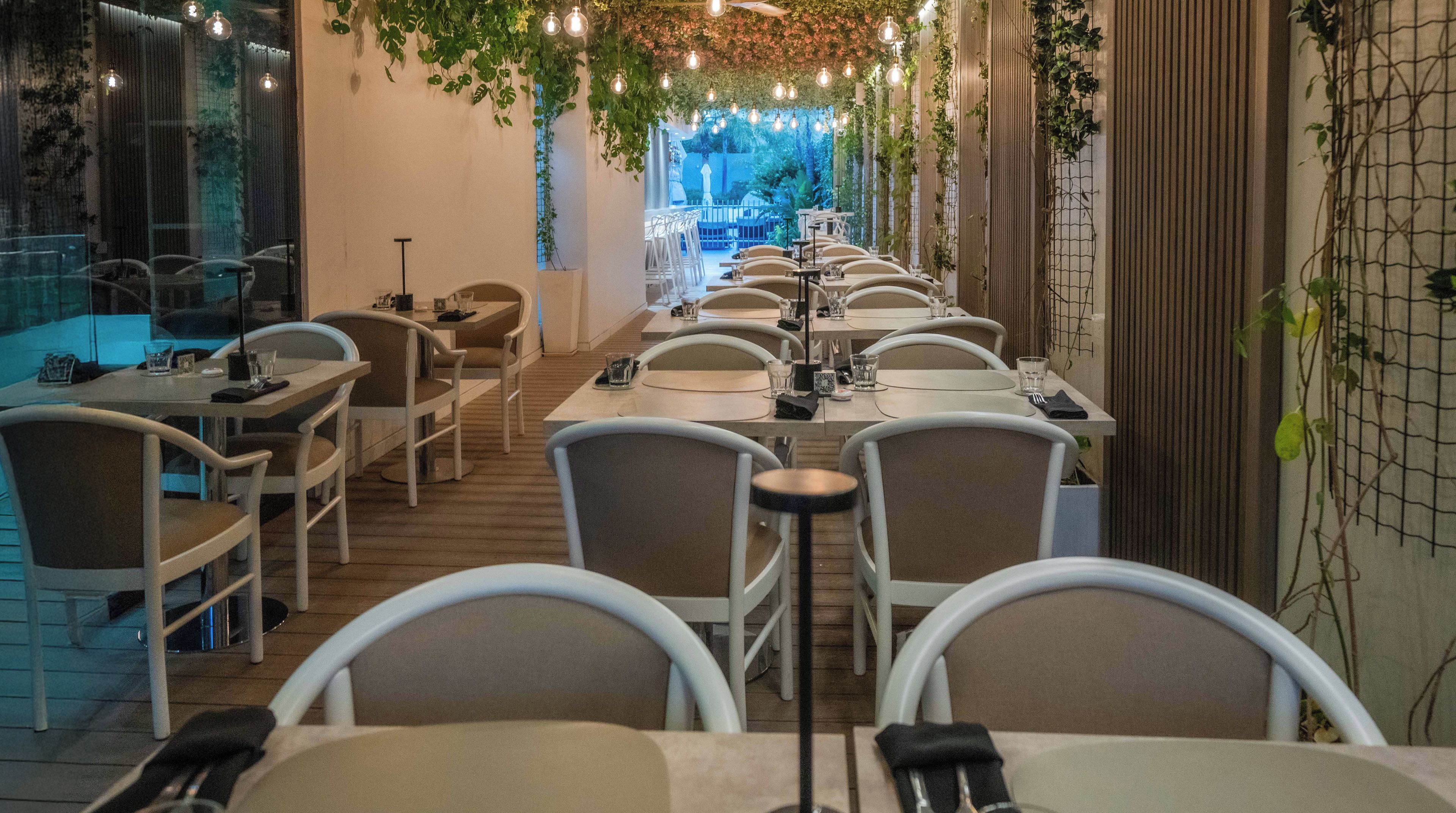 A restaurant with long tables and chairs, hanging lights, and greenery.