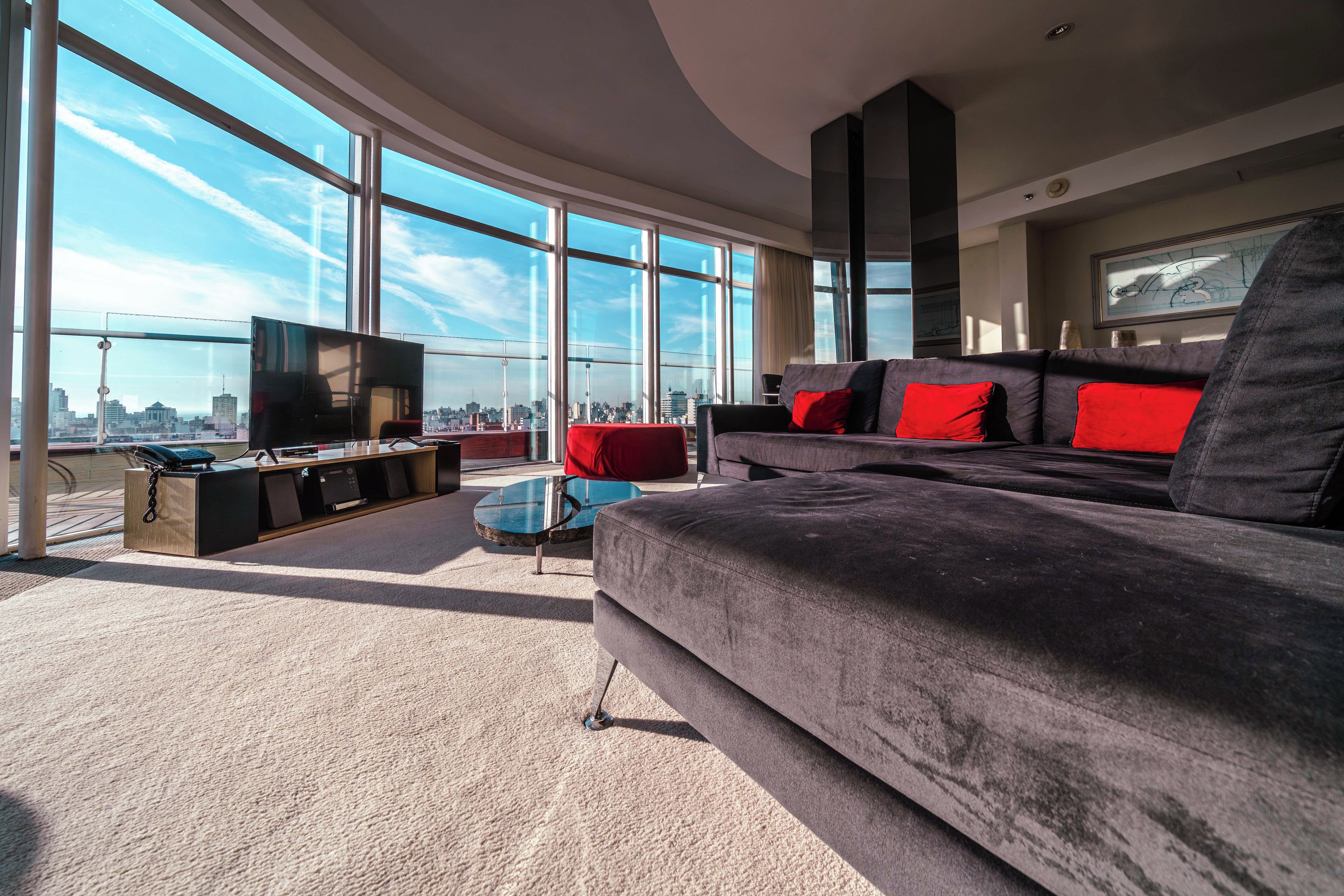 Een moderne woonkamer met een groot raam, een flatscreen televisie, een donkere bank met rode kussens en een glazen salontafel.