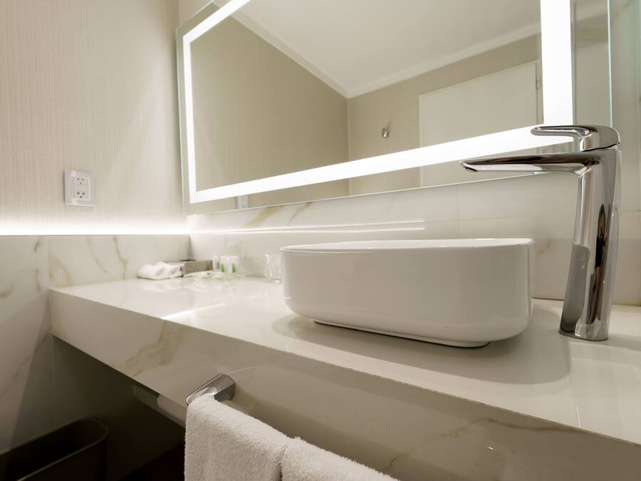 A bathroom with a white marble countertop, a white sink, a chrome faucet, and a mirror with a light above it.