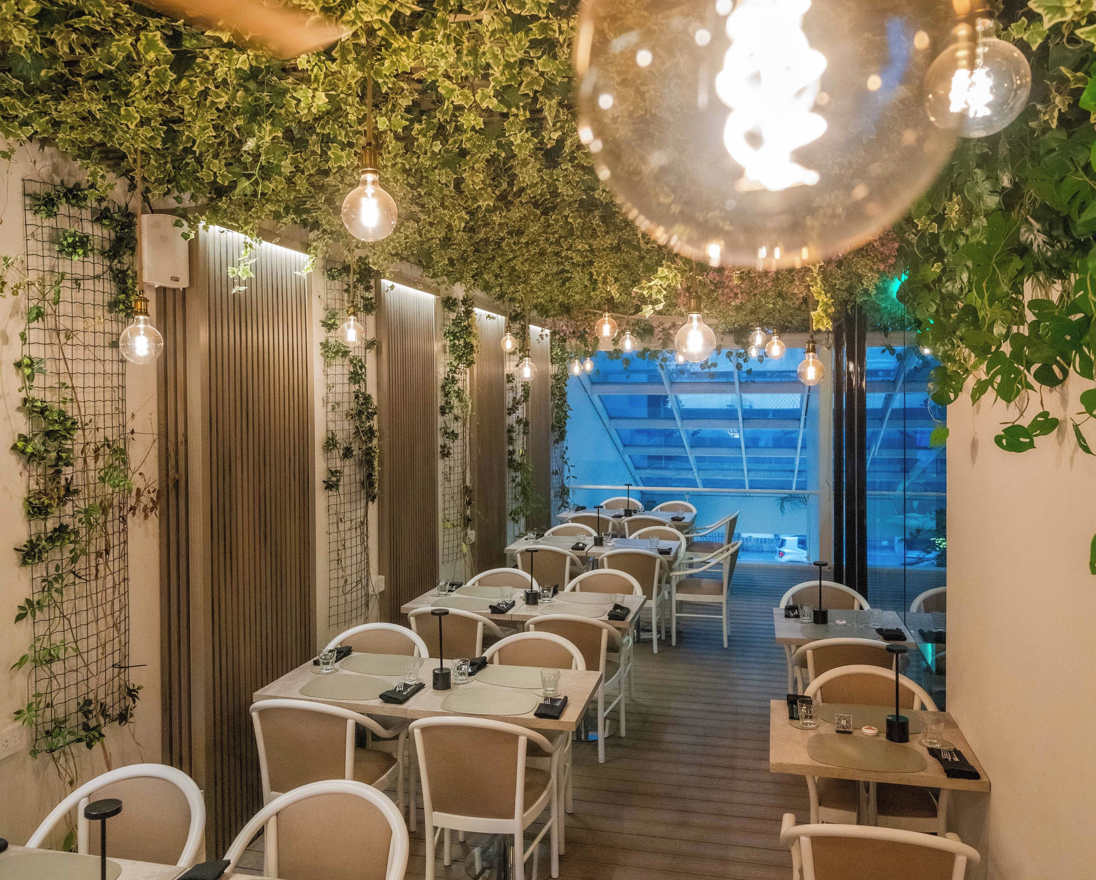 Un restaurante con mesas y sillas dispuestas en filas, un techo con luces colgantes y paredes cubiertas de plantas trepadoras.