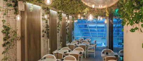 Un restaurante con mesas y sillas dispuestas en filas, un techo con luces colgantes y paredes cubiertas de plantas trepadoras.