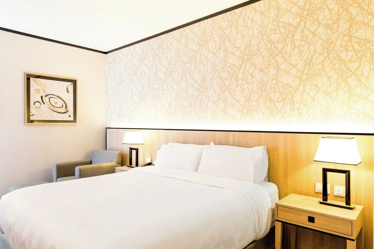 Een hotelkamer met een groot bed, twee nachtkastjes met lampen, een stoel en een kunstwerk aan de muur.