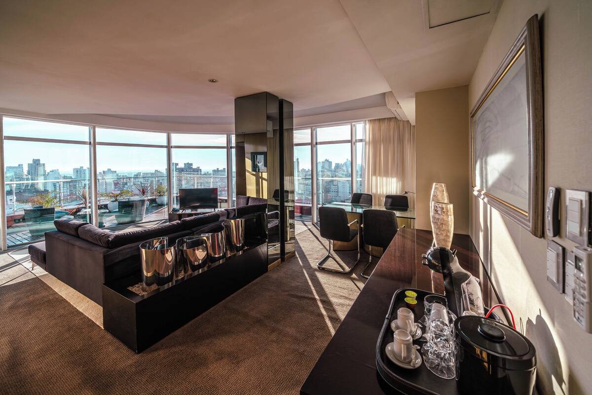 A modern hotel room with a large window offering a city view, a black sofa, a coffee station, and a desk with chairs.