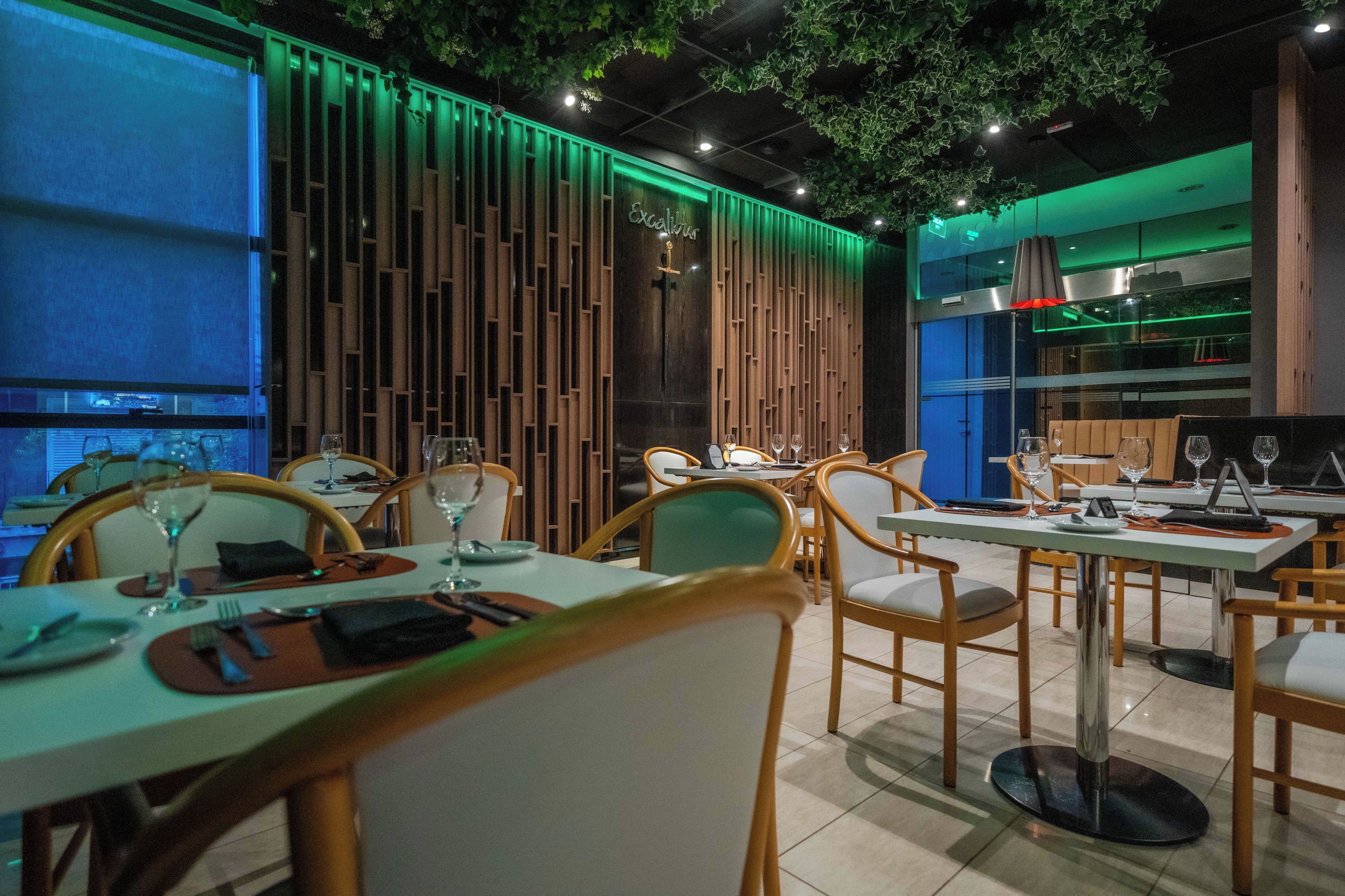 A restaurant with wooden panel walls, green ambient lighting, and a modern kitchen visible through a glass partition.