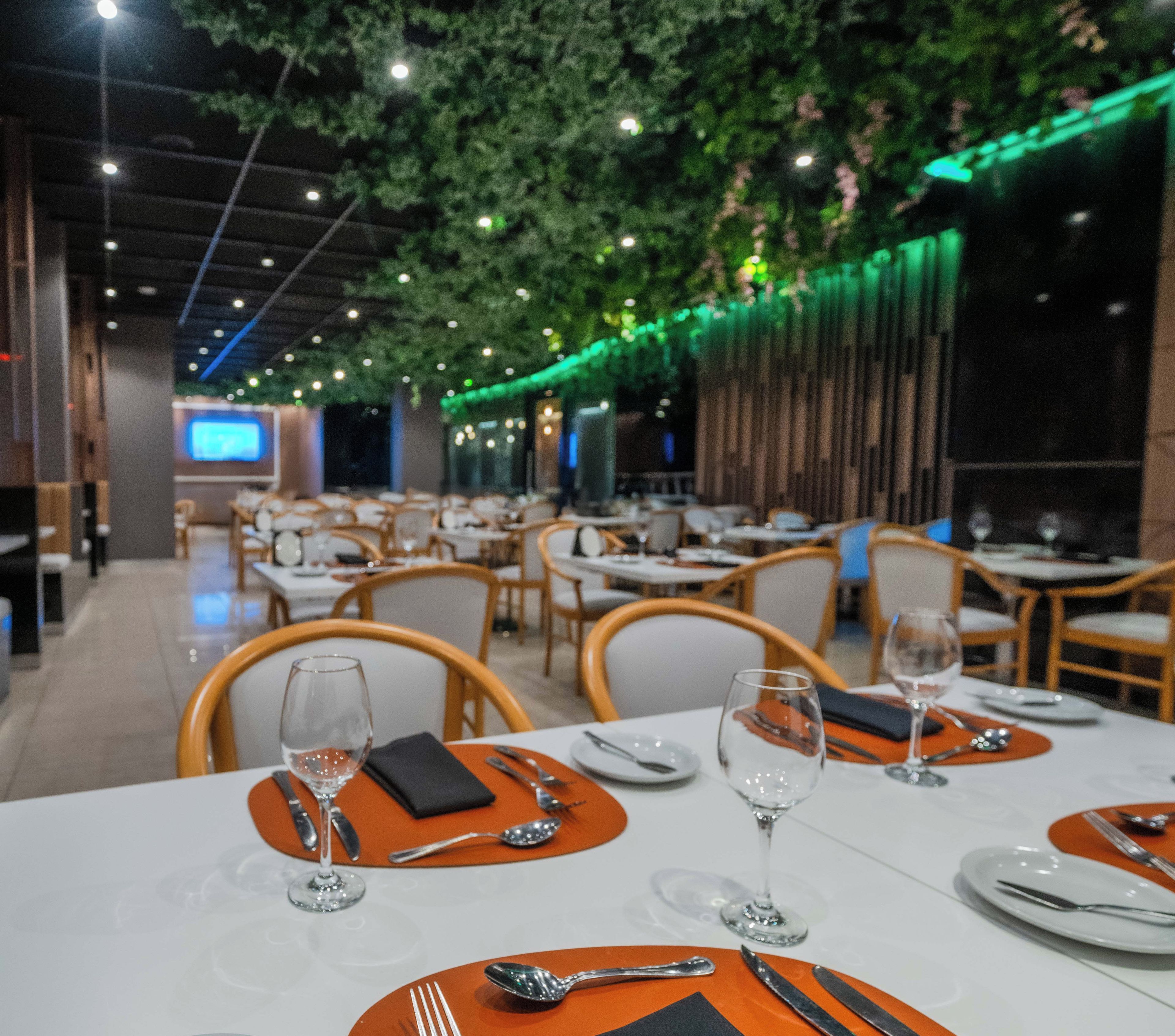 A restaurant with tables set for dining, featuring a modern interior with green foliage hanging from the ceiling and ambient lighting.