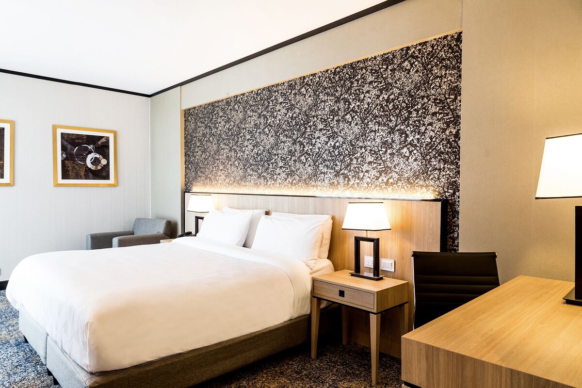 A hotel room with a large bed, two framed pictures on the wall, a desk with a chair, and a large abstract painting above the bed.