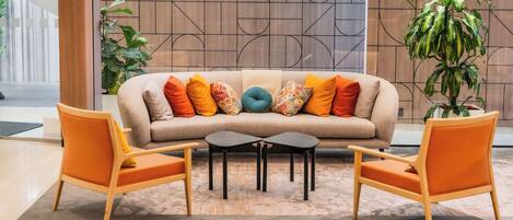 Un área de descanso moderna con un sofá color beige, cojines coloridos, dos sillones de madera y una mesita de centro.