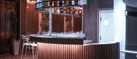 Zona de bar con mostrador de madera, varias estanterías llenas de botellas y dos taburetes blancos.