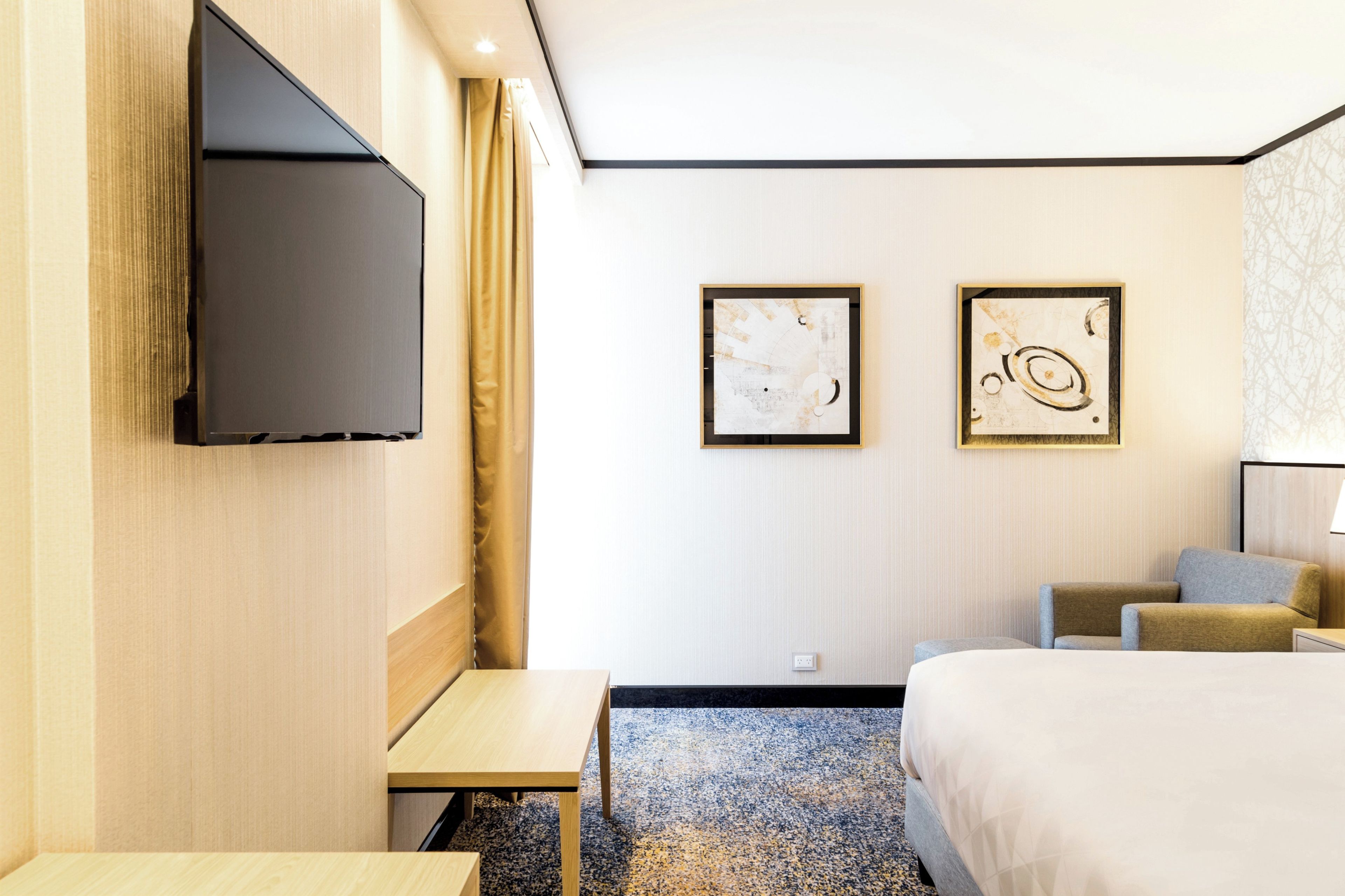 Een hotelkamer met een bed, een bureau, een stoel, twee ingelijste kunstwerken aan de muur en een aan de muur bevestigde televisie.