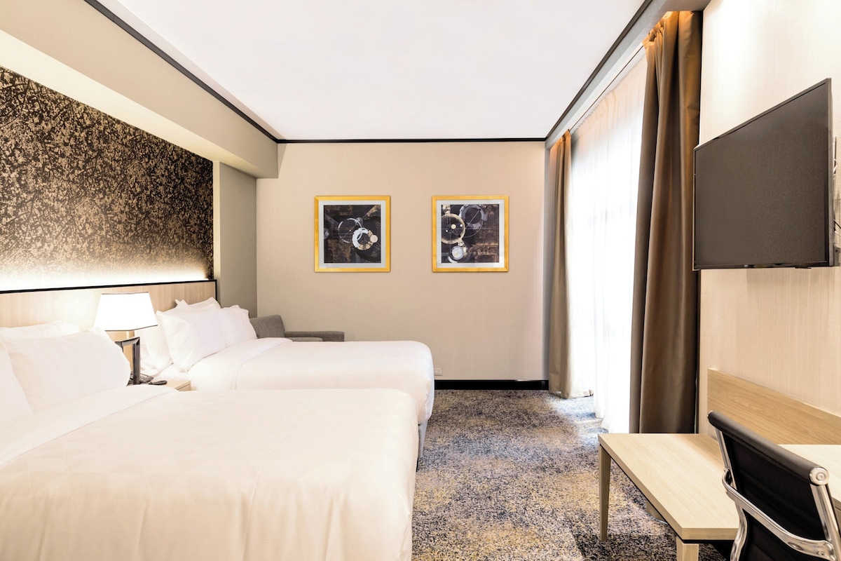 Une chambre d’hôtel avec deux lits, un bureau, une chaise, une télévision et des œuvres d’art accrochées au mur.