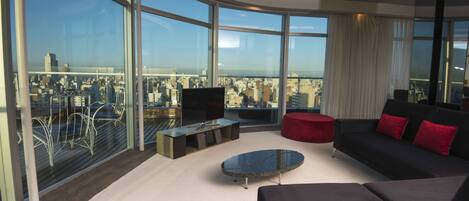 Una sala moderna con un amplio ventanal, un televisor de pantalla plana, un sofá, un reposapiés rojo y una mesa de centro.