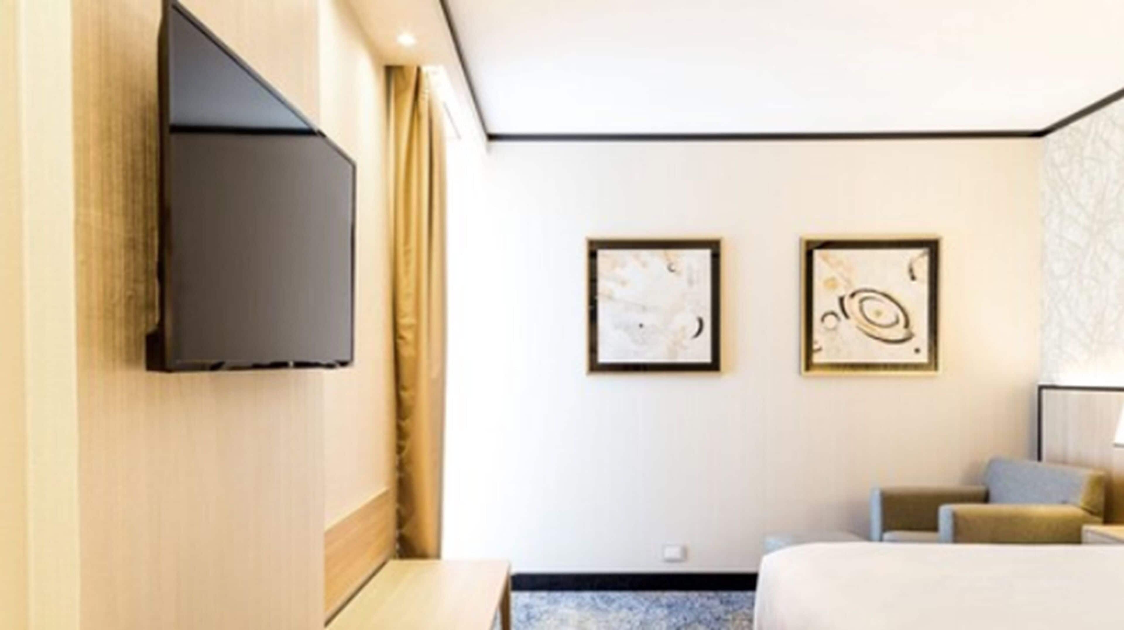 Quarto de hotel com televisão de tela plana na parede, duas pinturas abstratas emolduradas, cama com cabeceira azul e uma pequena mesa.