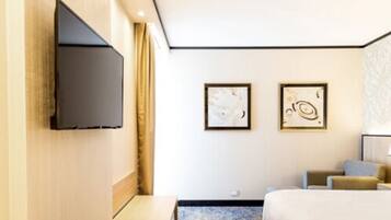 Quarto de hotel com televisão de tela plana na parede, duas pinturas abstratas emolduradas, cama com cabeceira azul e uma pequena mesa.