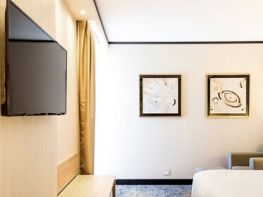 A hotel room with a flat-screen TV mounted on the wall, two framed abstract paintings, a bed with a blue headboard, and a small table.