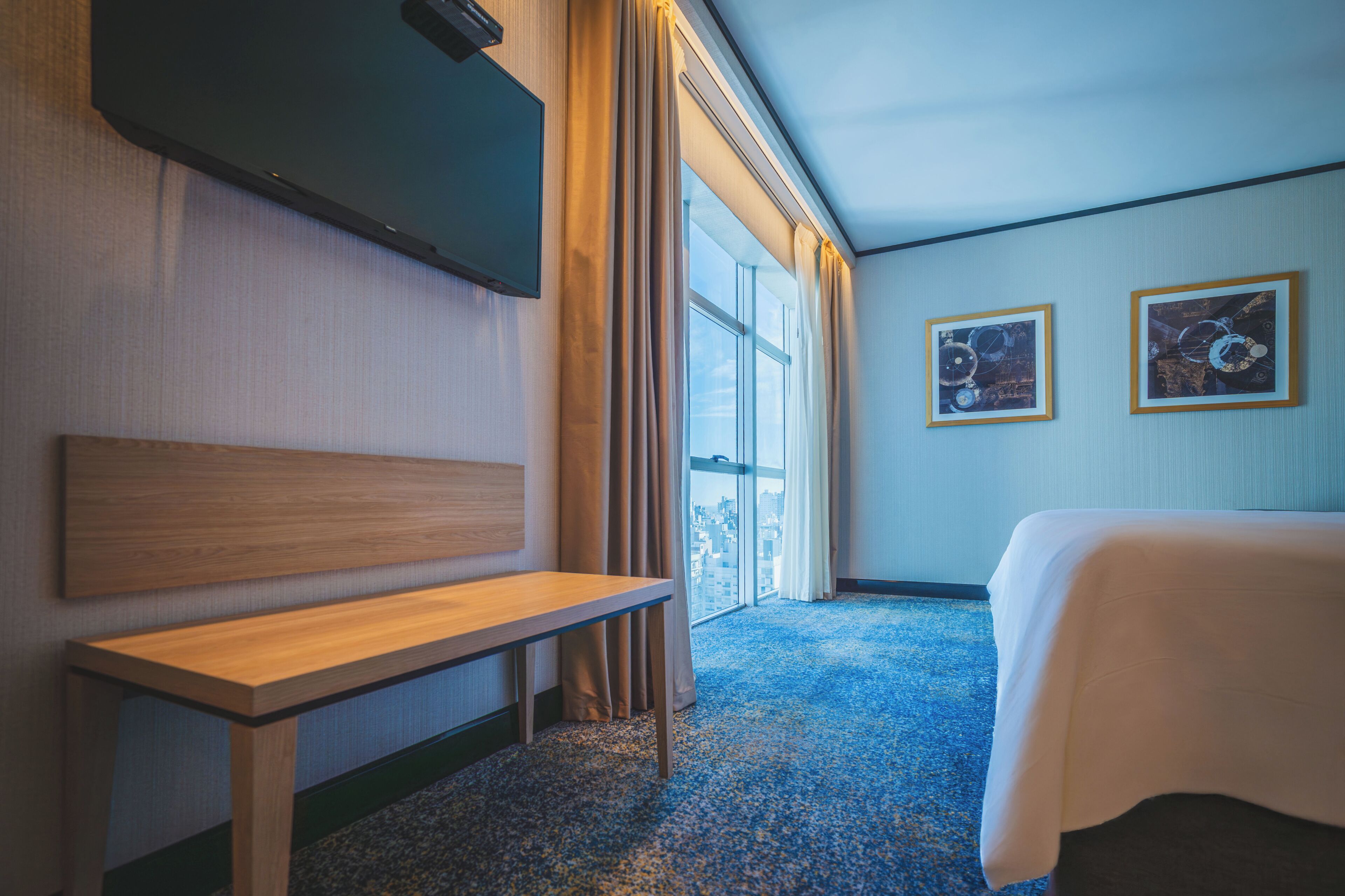 Habitación de hotel con cama, un banco, televisor y dos cuadros en la pared.
