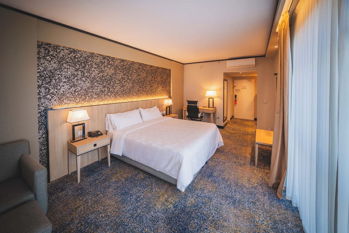 A hotel room with a bed, bedside tables, a chair, a desk, and a large wall art piece.