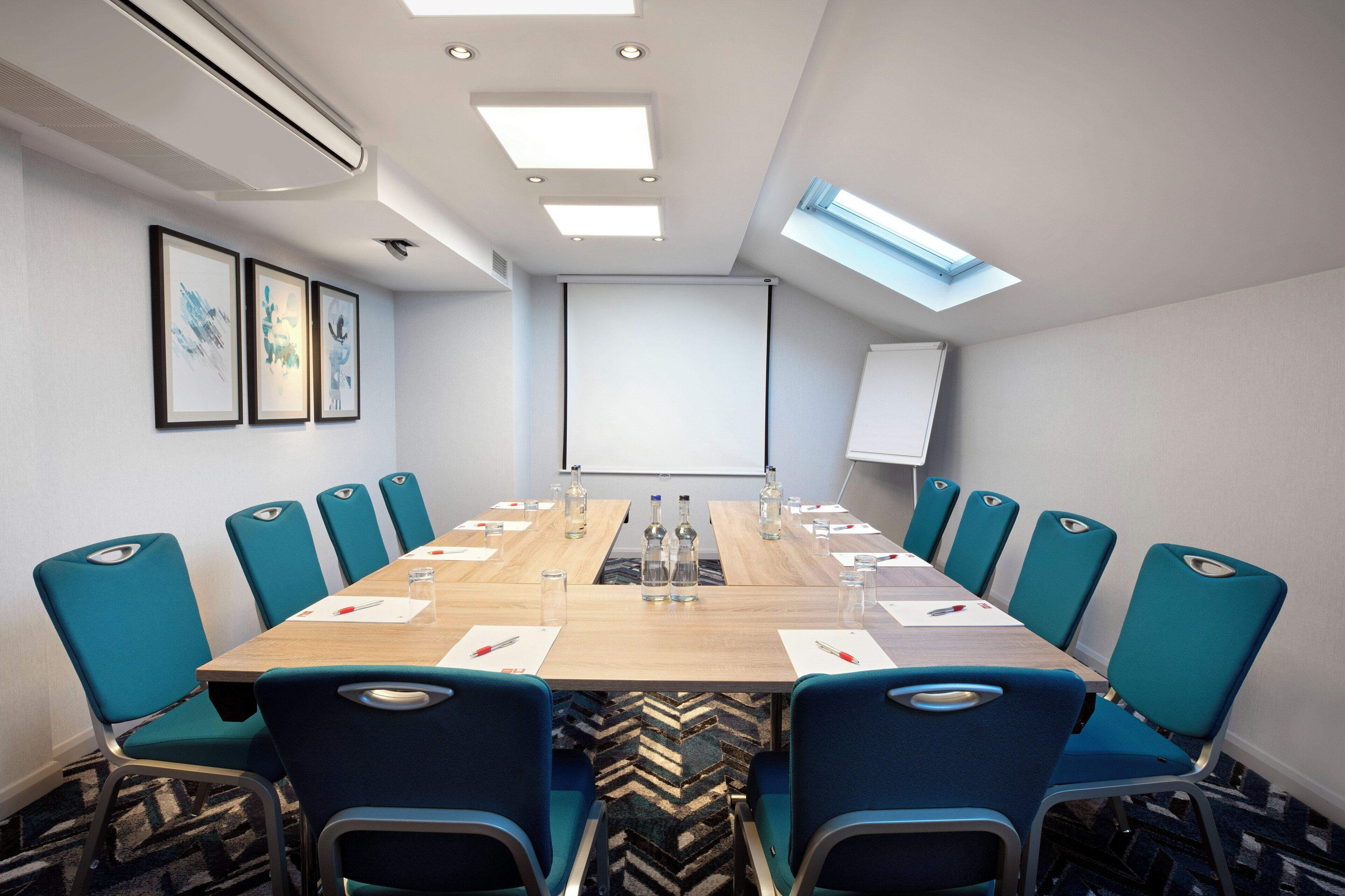 A conference room with a long table, teal chairs, and a projector screen.