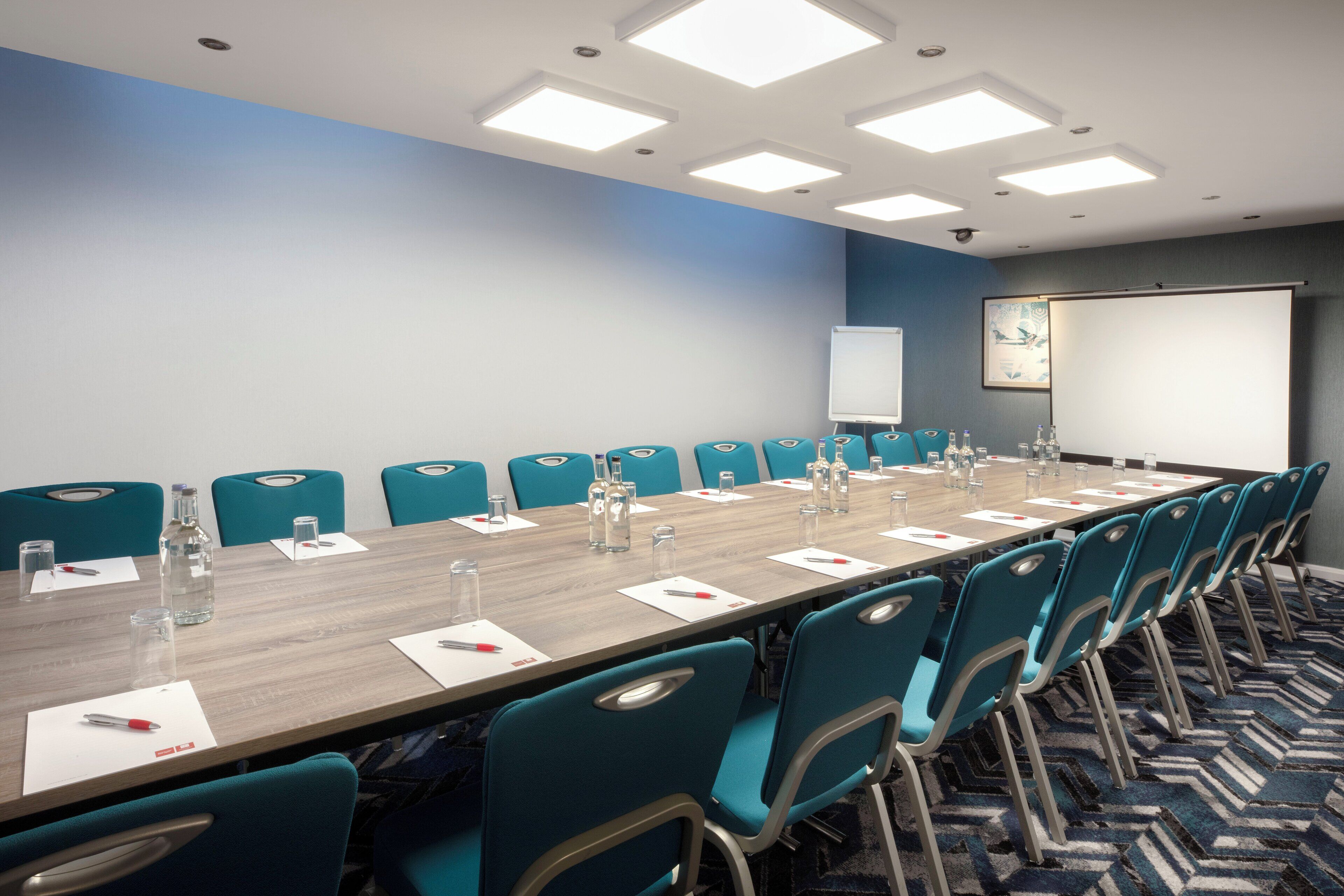 Un salón de conferencias con una larga mesa, sillas color turquesa y una pantalla para proyector.