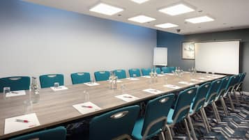 Un salón de conferencias con una larga mesa, sillas color turquesa y una pantalla para proyector.