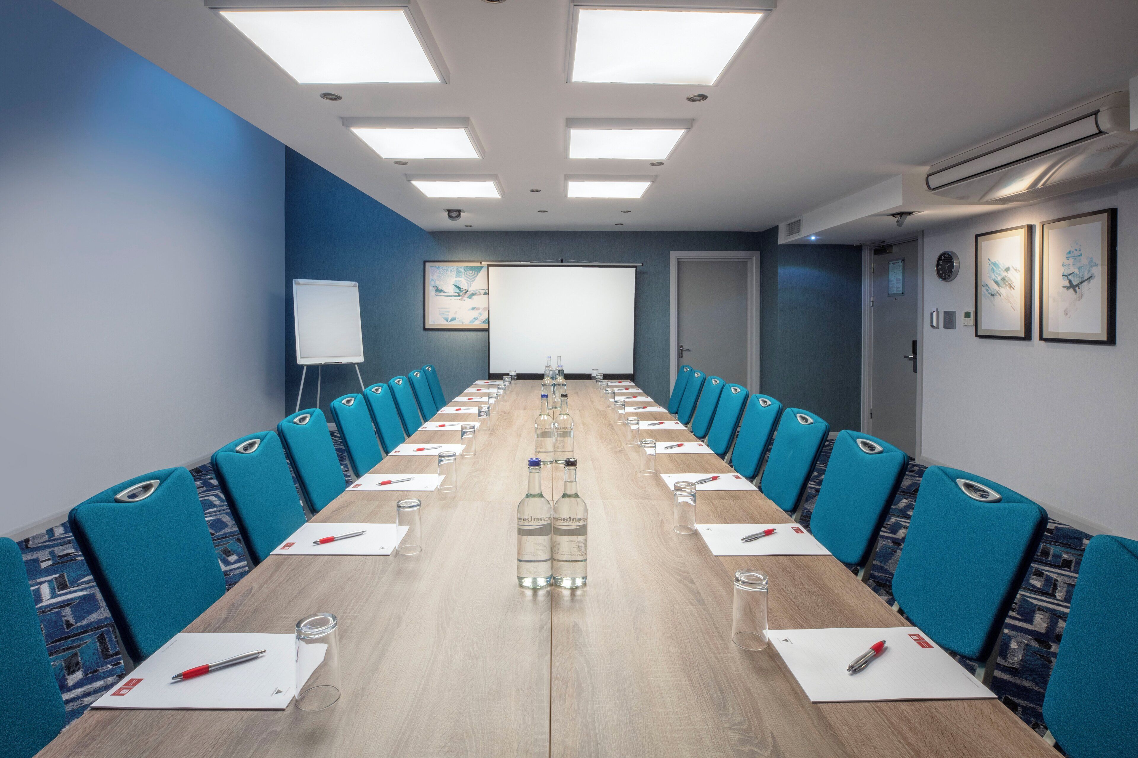 Ein Konferenzraum mit einem langen Tisch, blauen Stühlen und einer Whiteboard-Präsentation an der Wand.