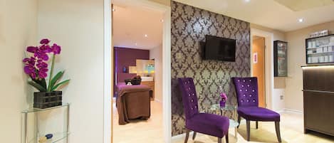 Hall d'albergo con sedie viola, un tavolo in vetro, una televisione a parete e una reception sullo sfondo.