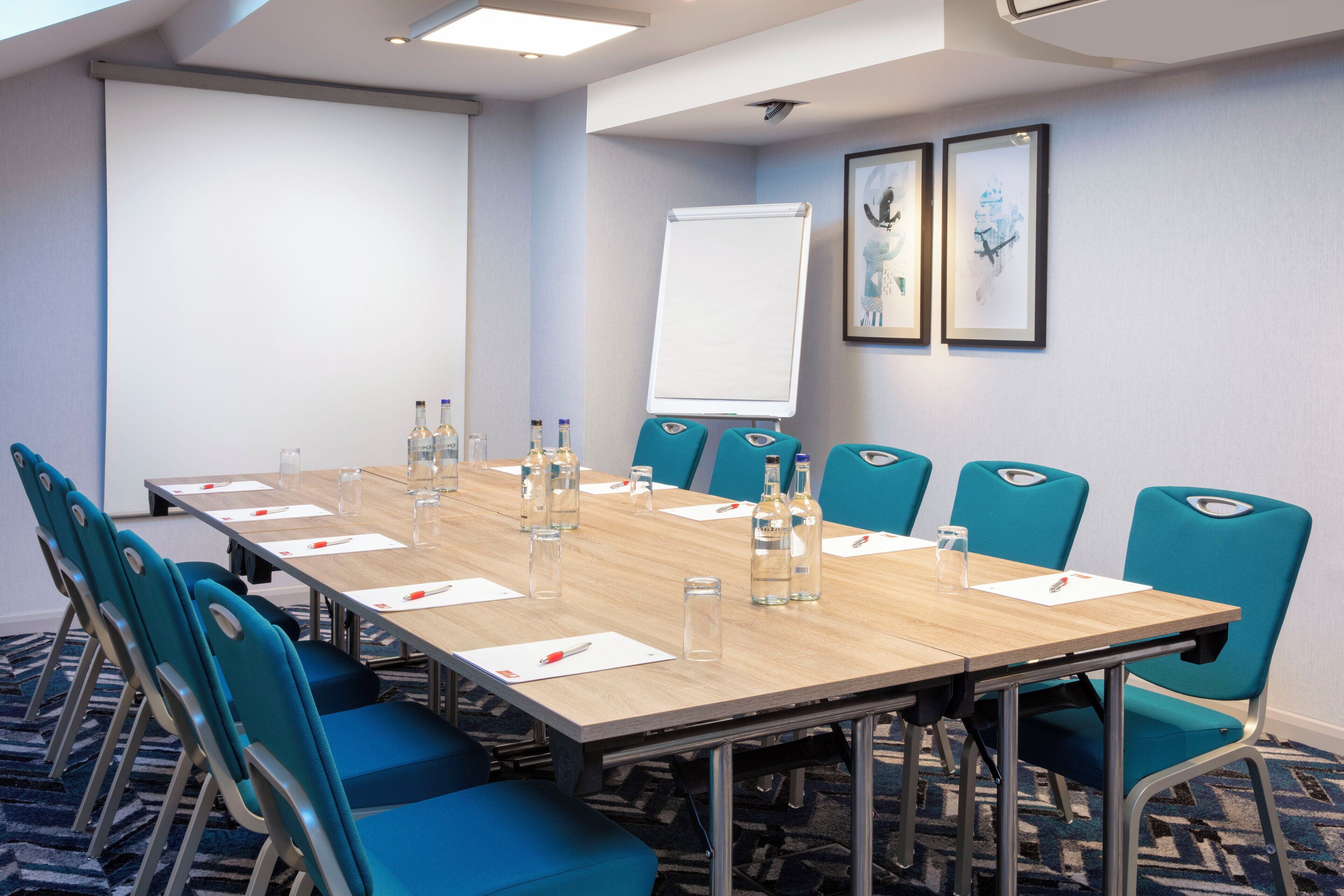 A conference room with a long table, blue chairs, a whiteboard, and framed artwork on the wall.