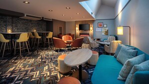 En moderne hotellobby med højt til loftet, et mønstret tæppe, en blå sofa, orange stole og et barområde.