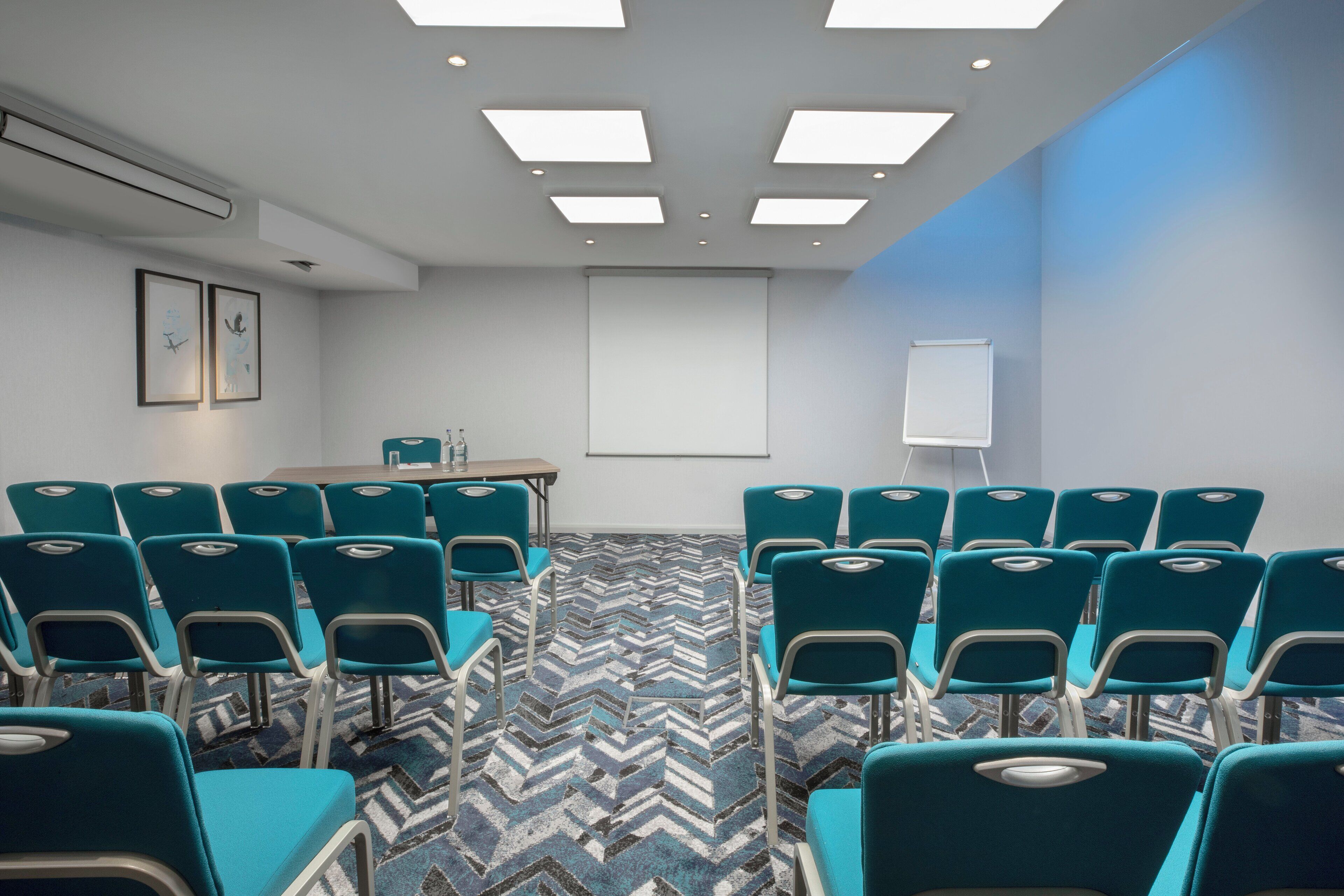 A conference room with teal chairs, a projector screen, and a patterned carpet.