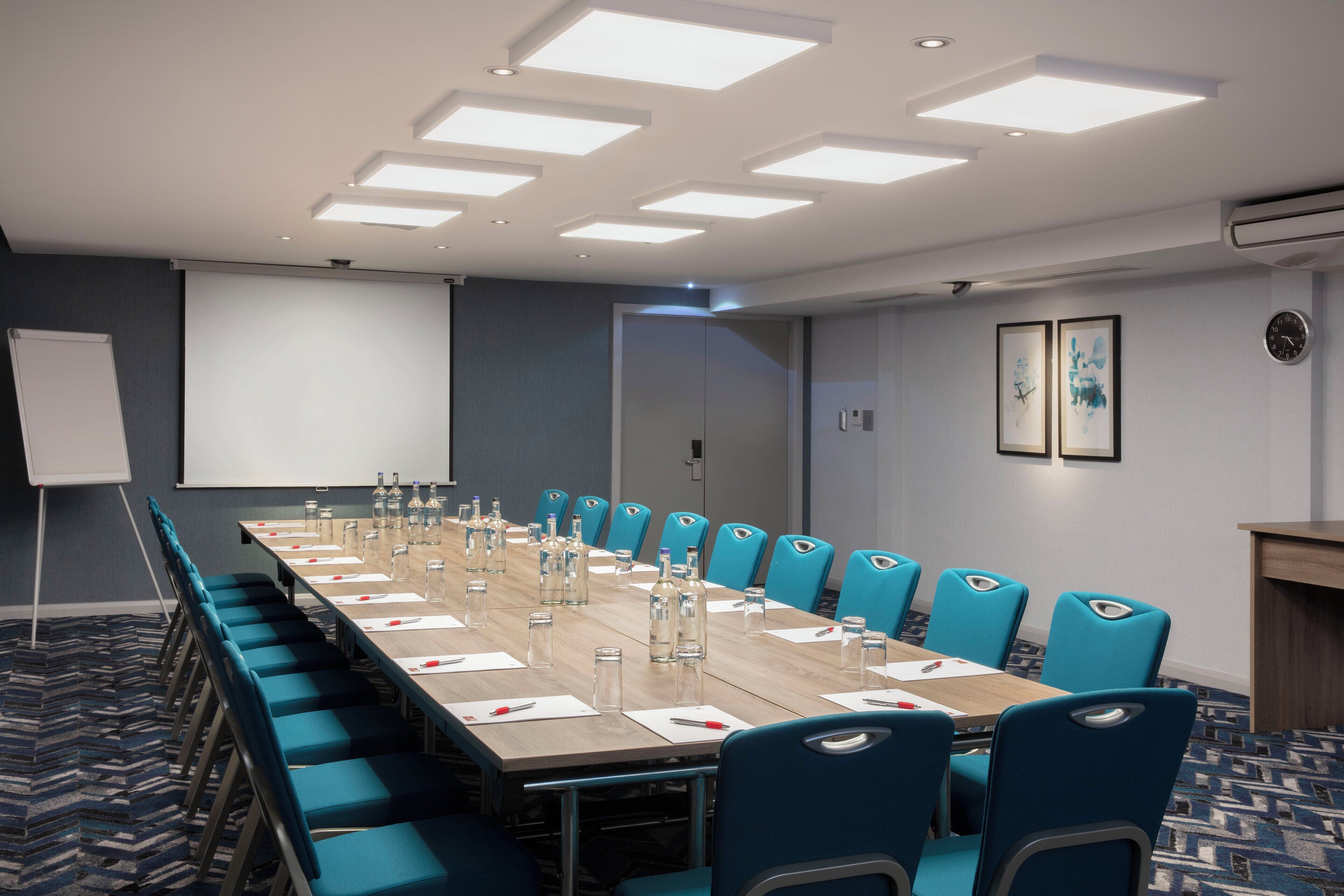 Un salón de conferencias con una larga mesa, sillas azules y una pantalla de proyector.