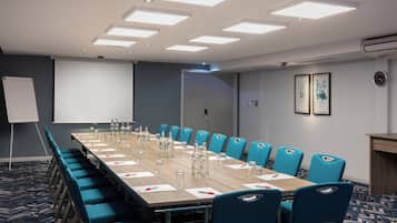 Un salón de conferencias con una larga mesa, sillas azules y una pantalla de proyector.