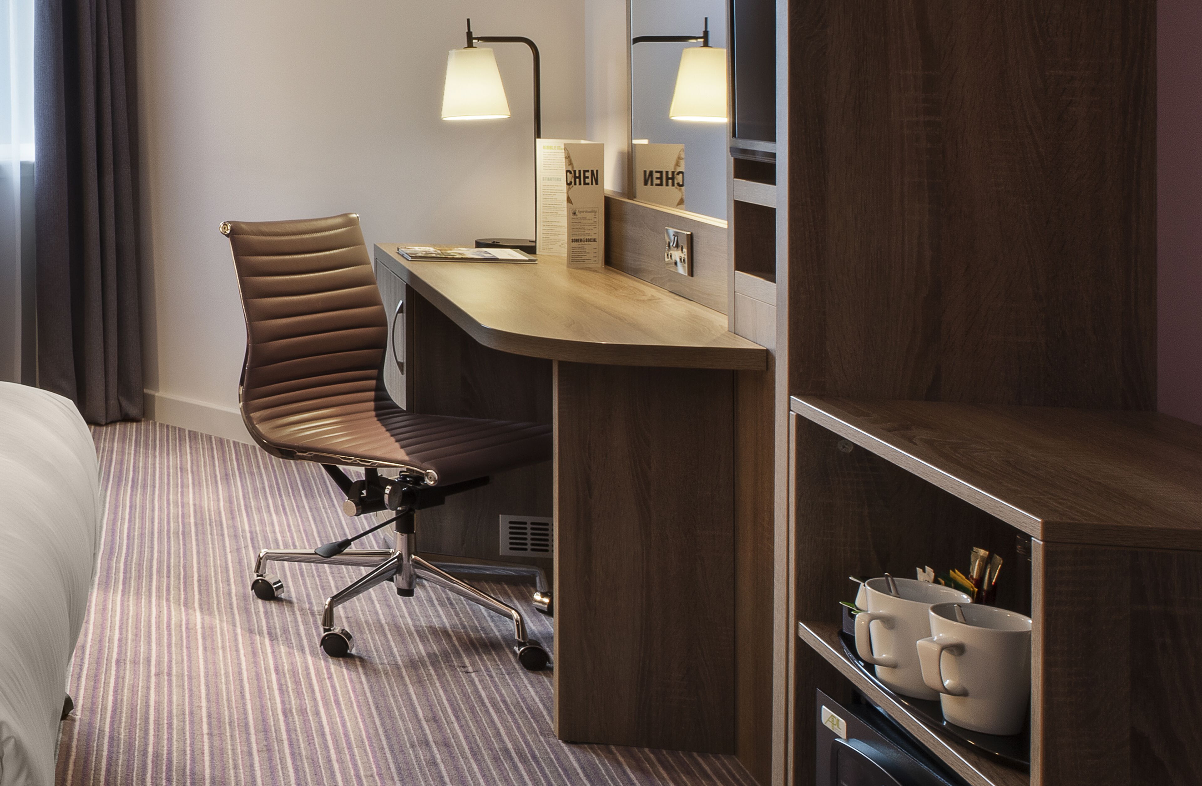 A hotel room with a wooden desk, a cushioned chair, a bed, and a shelf with mugs.