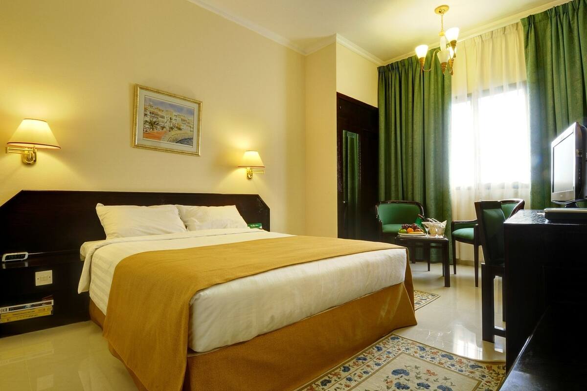 A hotel room with a bed, two bedside lamps, a desk with a computer, a chair, and a window with curtains.