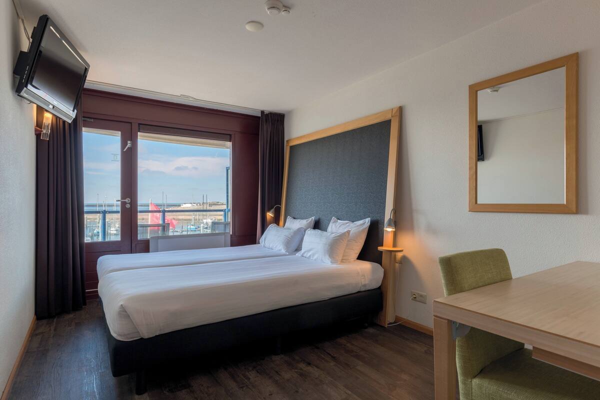 Una habitación de hotel con cama, escritorio, silla, televisor y ventana con cortinas.
