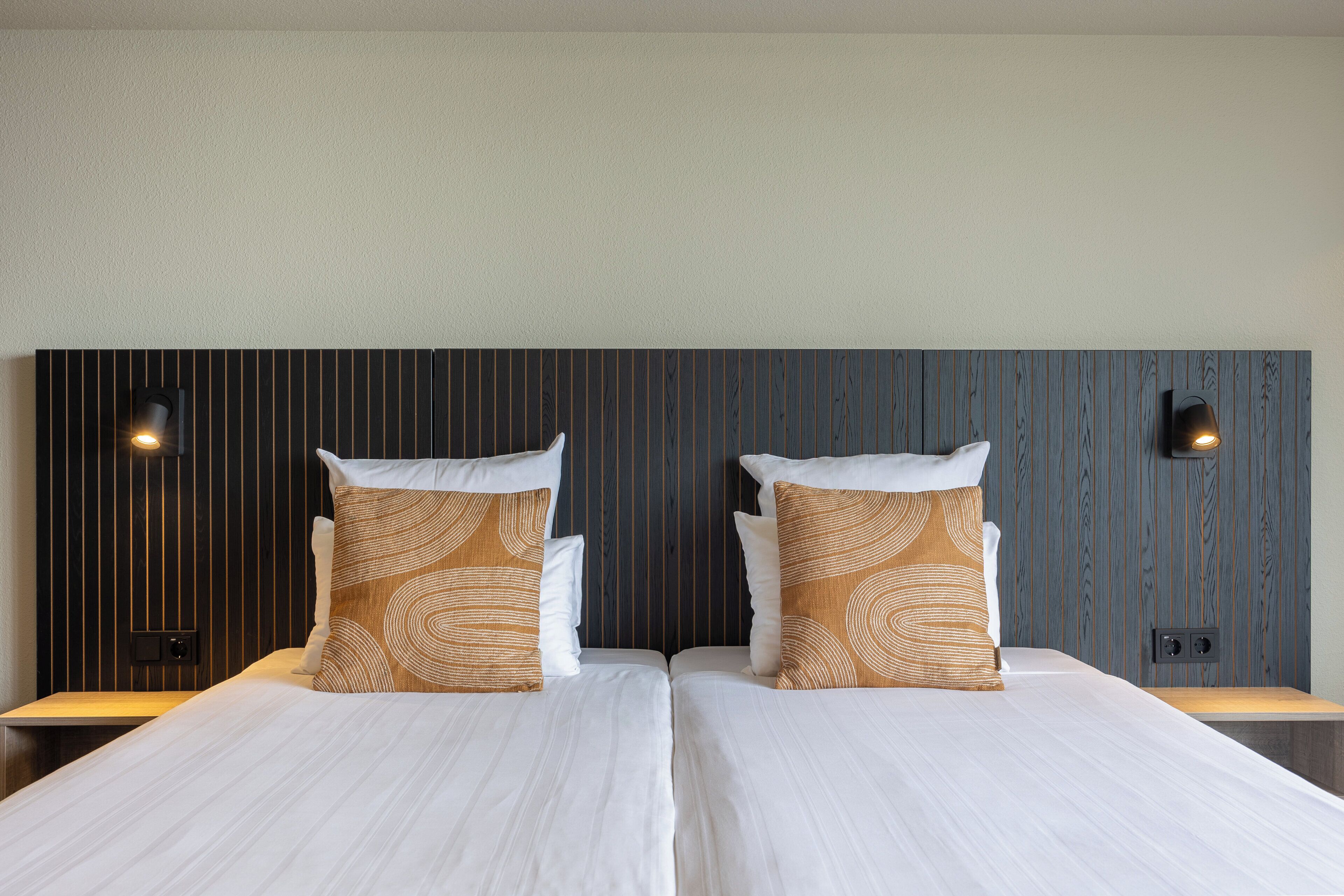 A hotel room with two beds, each with white linens and patterned pillows, a dark wood panel headboard, and wall-mounted reading lights.
