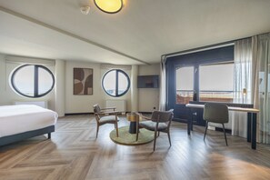 Une chambre d’hôtel avec un lit, deux chaises, une petite table, une fenêtre ronde et une grande fenêtre avec des rideaux.