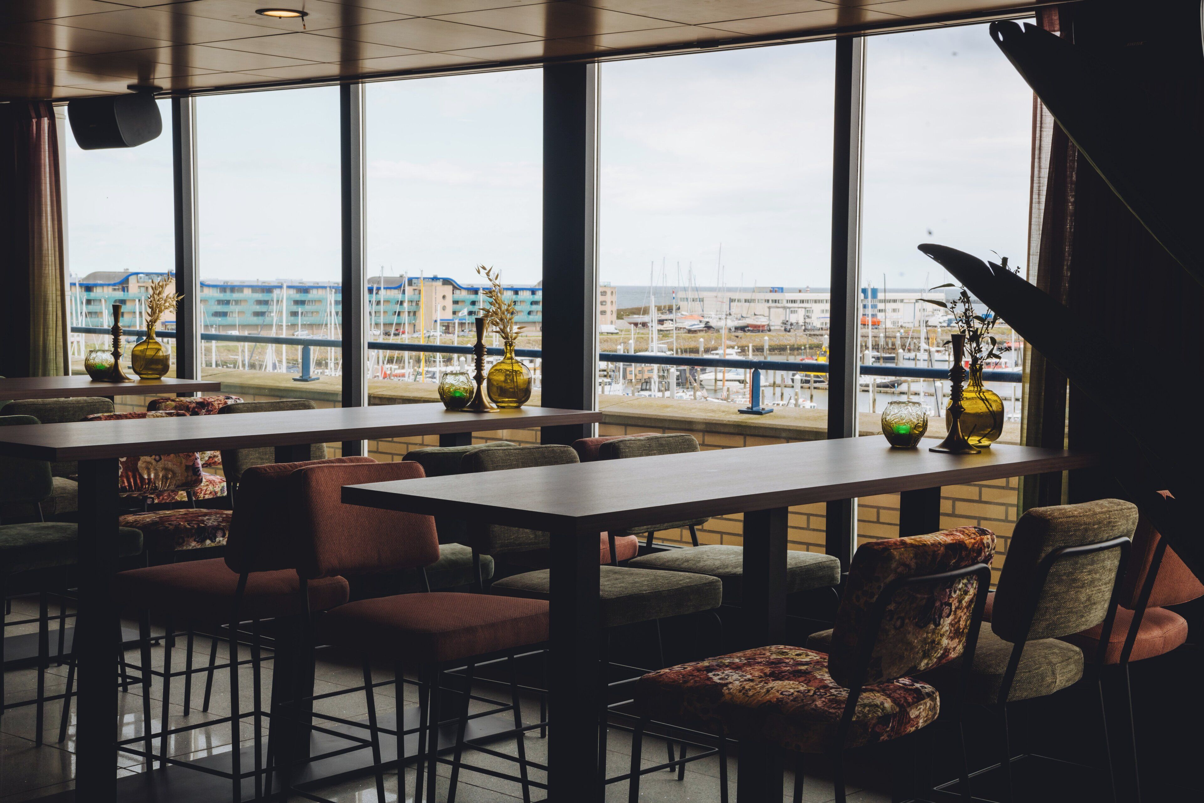 Un restaurant doté de grandes fenêtres offrant une vue sur un port de plaisance et des bâtiments. Des tables et des chaises sont disposées pour accueillir les clients.