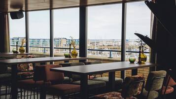 Un restaurant doté de grandes fenêtres offrant une vue sur un port de plaisance et des bâtiments. Des tables et des chaises sont disposées pour accueillir les clients.