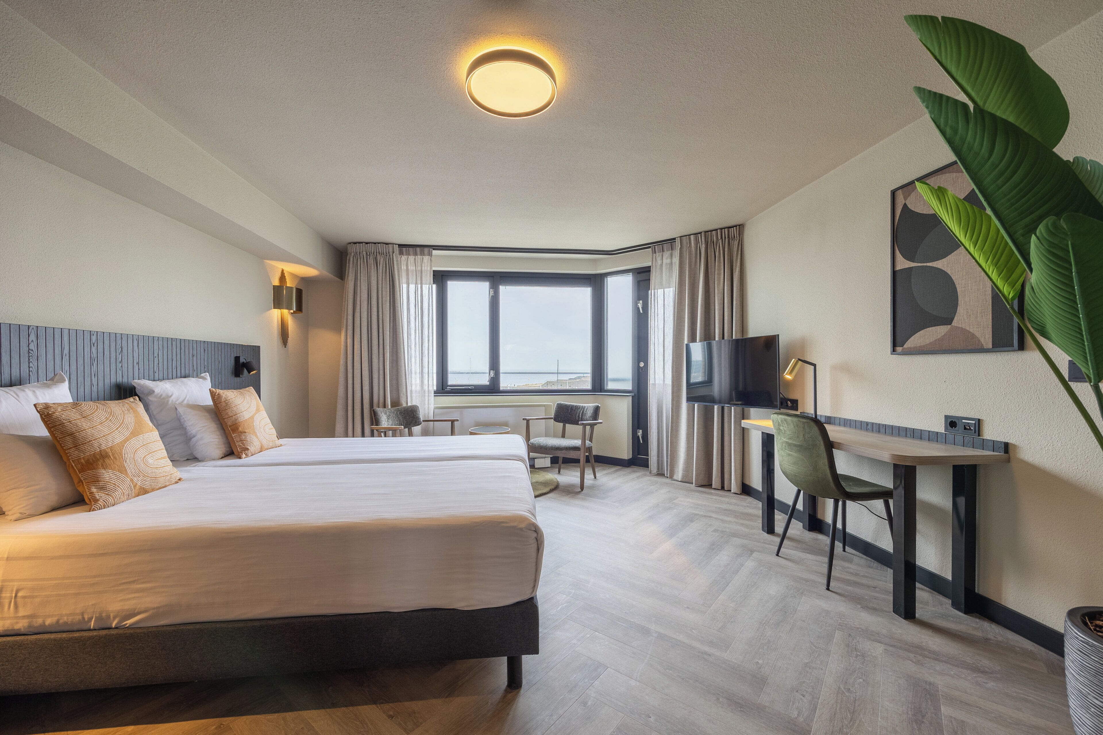 A hotel room with a large bed, a desk, a chair, and a TV. There is a window with curtains and a plant on the right side.