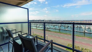 A balcony with a glass table and chairs, overlooking a marina with boats and a road.