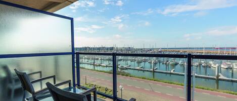 Un balcón con una mesa y sillas de vidrio, con vista a un puerto deportivo con barcos y una carretera.