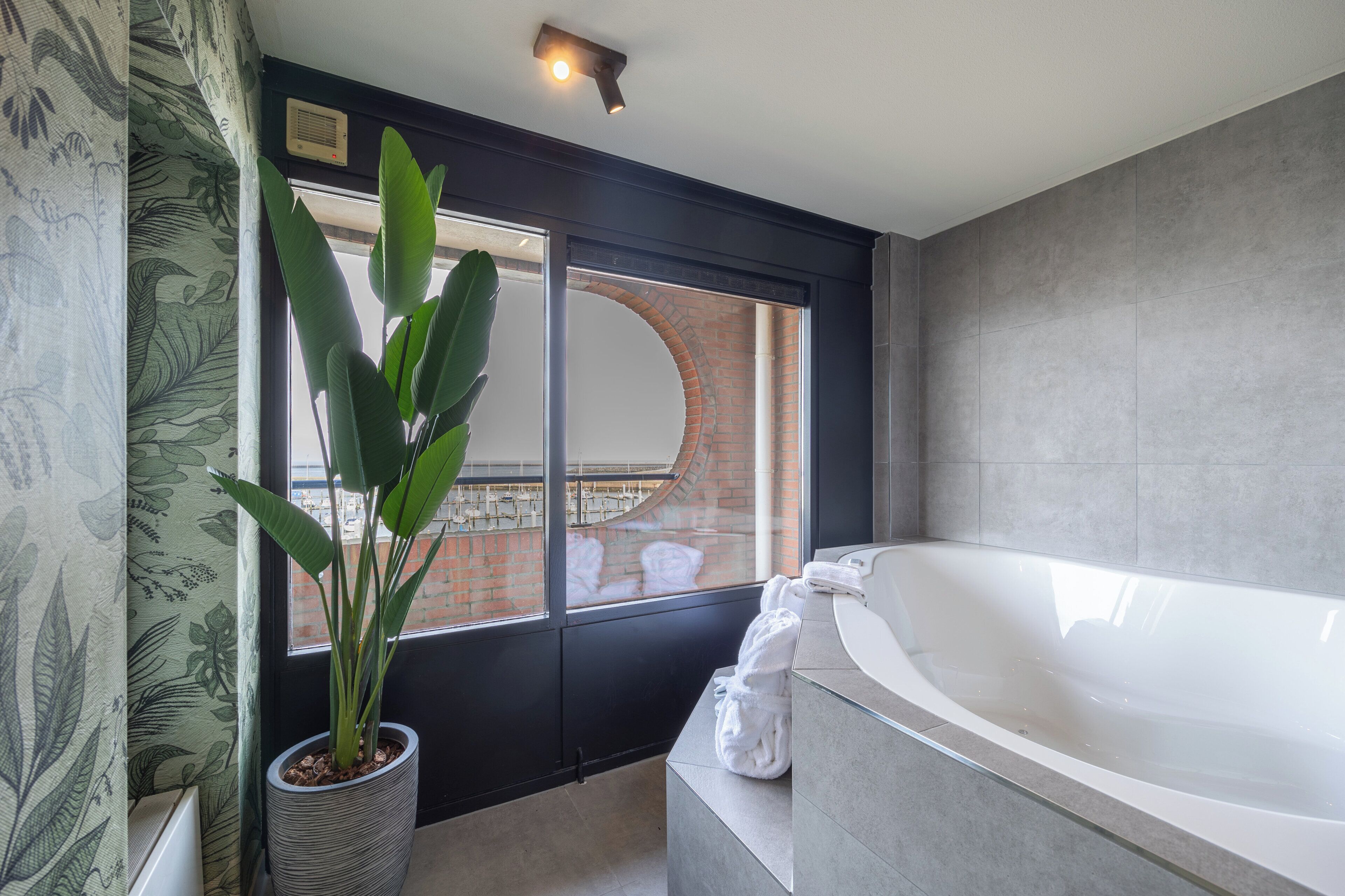 Ein modernes Badezimmer mit einer großen Badewanne, einem Topf mit Pflanze und Blick auf ein Ziegelgebäude durch ein großes Fenster.