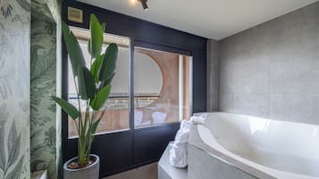 Ein modernes Badezimmer mit einer großen Badewanne, einem Topf mit Pflanze und Blick auf ein Ziegelgebäude durch ein großes Fenster.