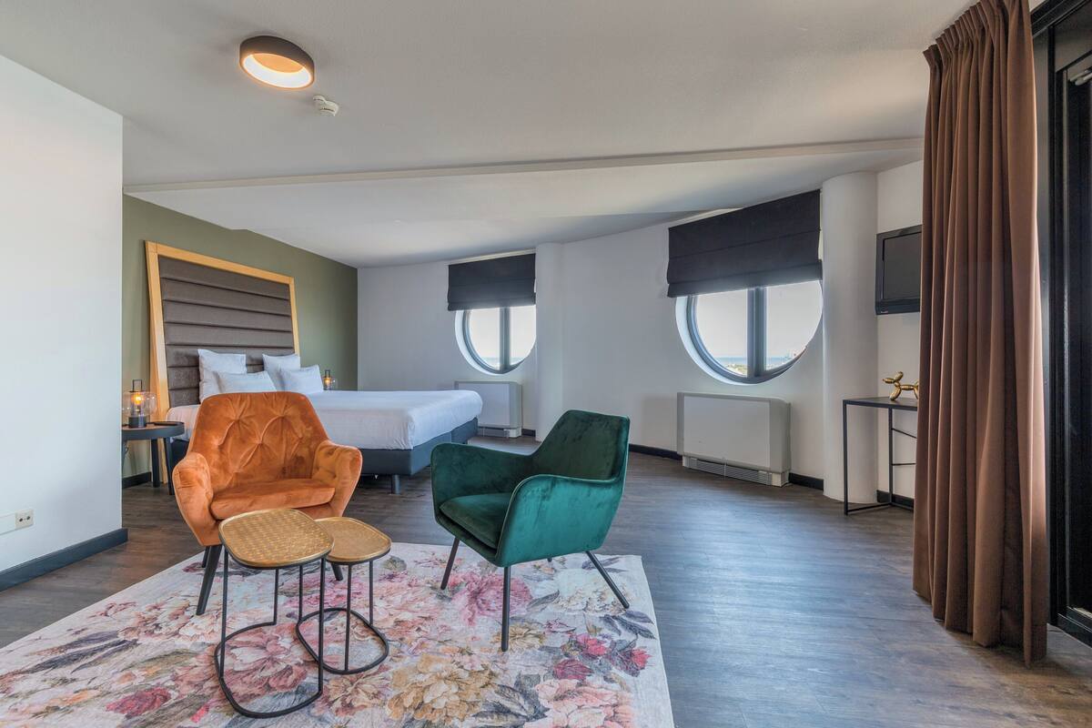Una habitación de hotel moderna con una cama, dos sillas, una mesita y un ventanal grande.
