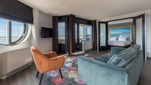 A modern hotel room with a large window, a sofa, a chair, a small table, and a bed with a view of the city.