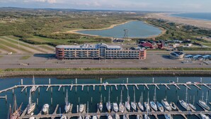 A marina with numerous boats docked, a large building with a distinctive roof, and a view of the surrounding landscape including a body of water and distant land.