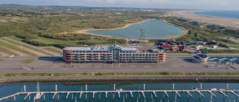 Un puerto deportivo con numerosas embarcaciones amarradas, un edificio grande con un techo distintivo y vistas del paisaje circundante, incluyendo una masa de agua y tierra en la distancia.