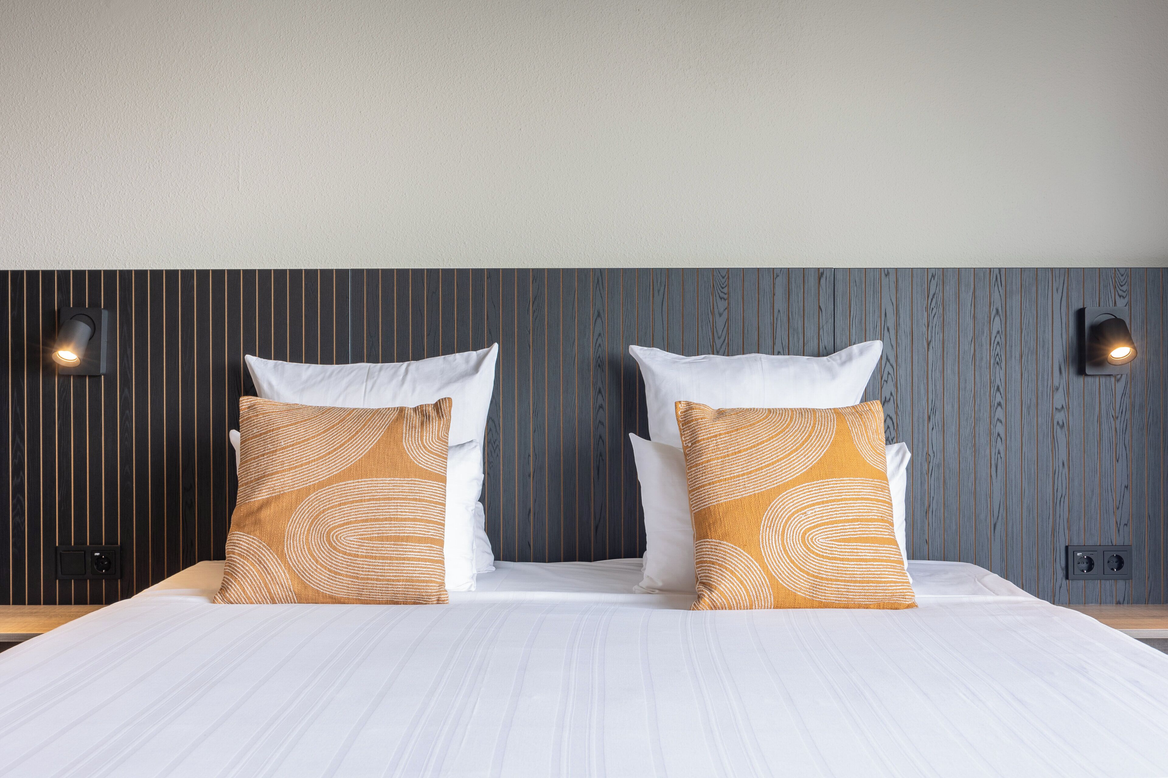 A neatly made bed with white linens and two decorative pillows, a wooden headboard, and two wall-mounted light fixtures.