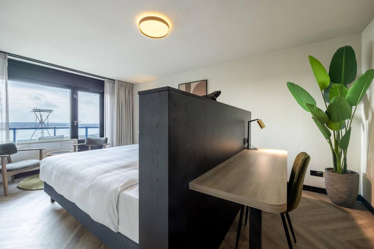 A hotel room with a bed, a desk, a chair, a potted plant, and a view of the sea.