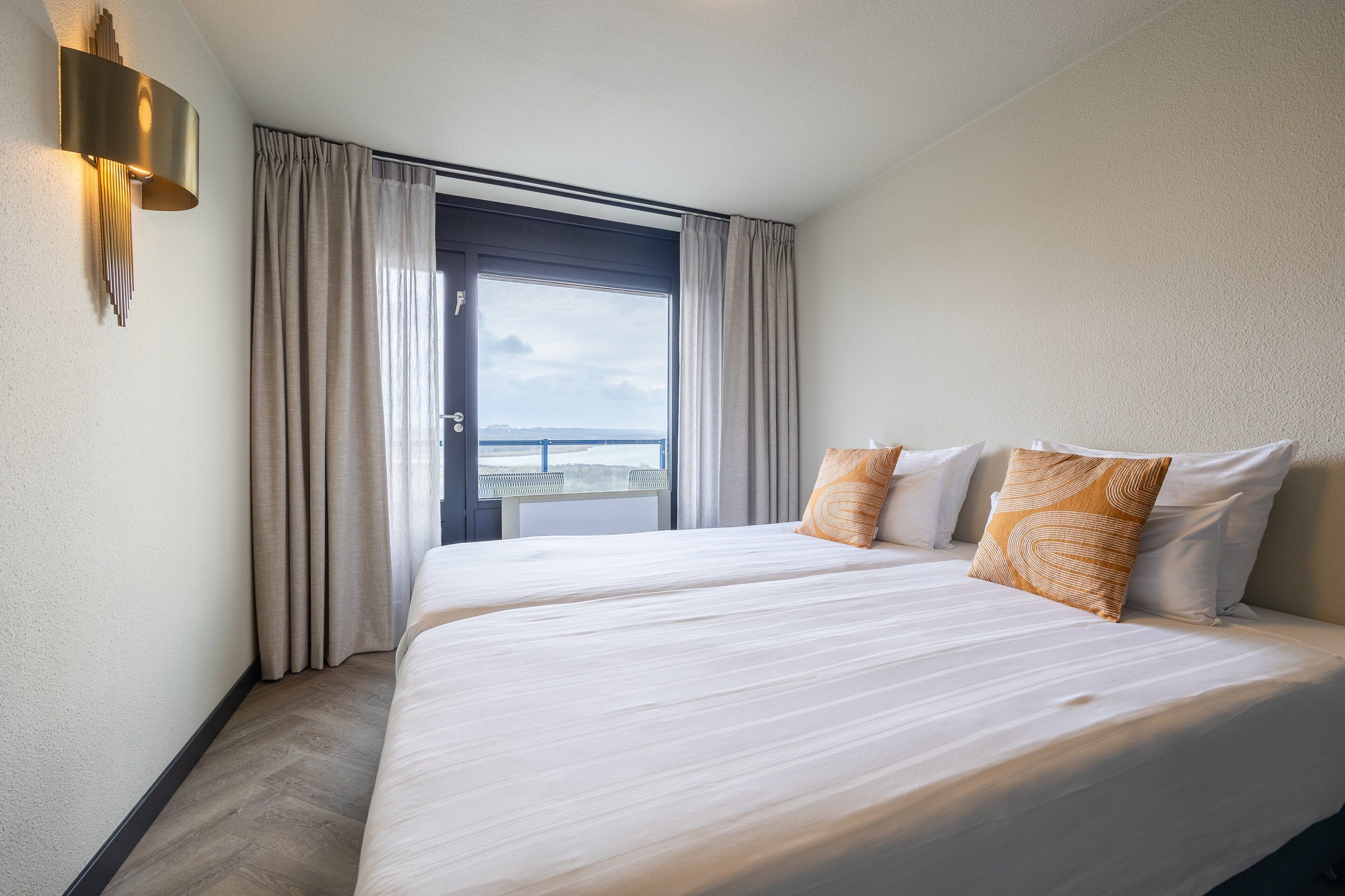 Una habitación de hotel con una cama grande, vistas al mar a través de una ventana y una lámpara moderna fijada a la pared.